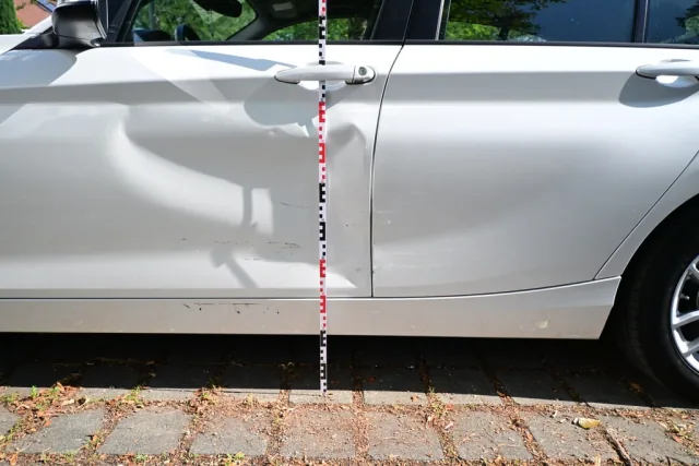 Weißer PKW mit eingedellter Seitenpartie, vermessen mit einer rot-weißen Messlatte.